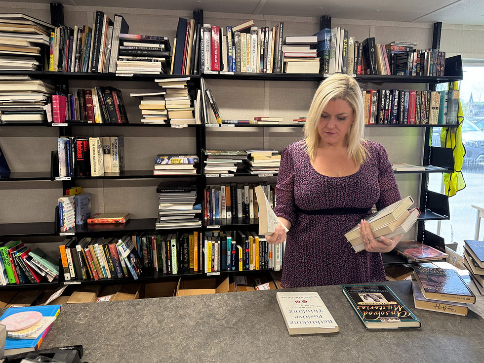 Westport Book Sale volunteer holds books, standing by a bookshelf. Books of various colors surround her. Sunlight peers through a window.