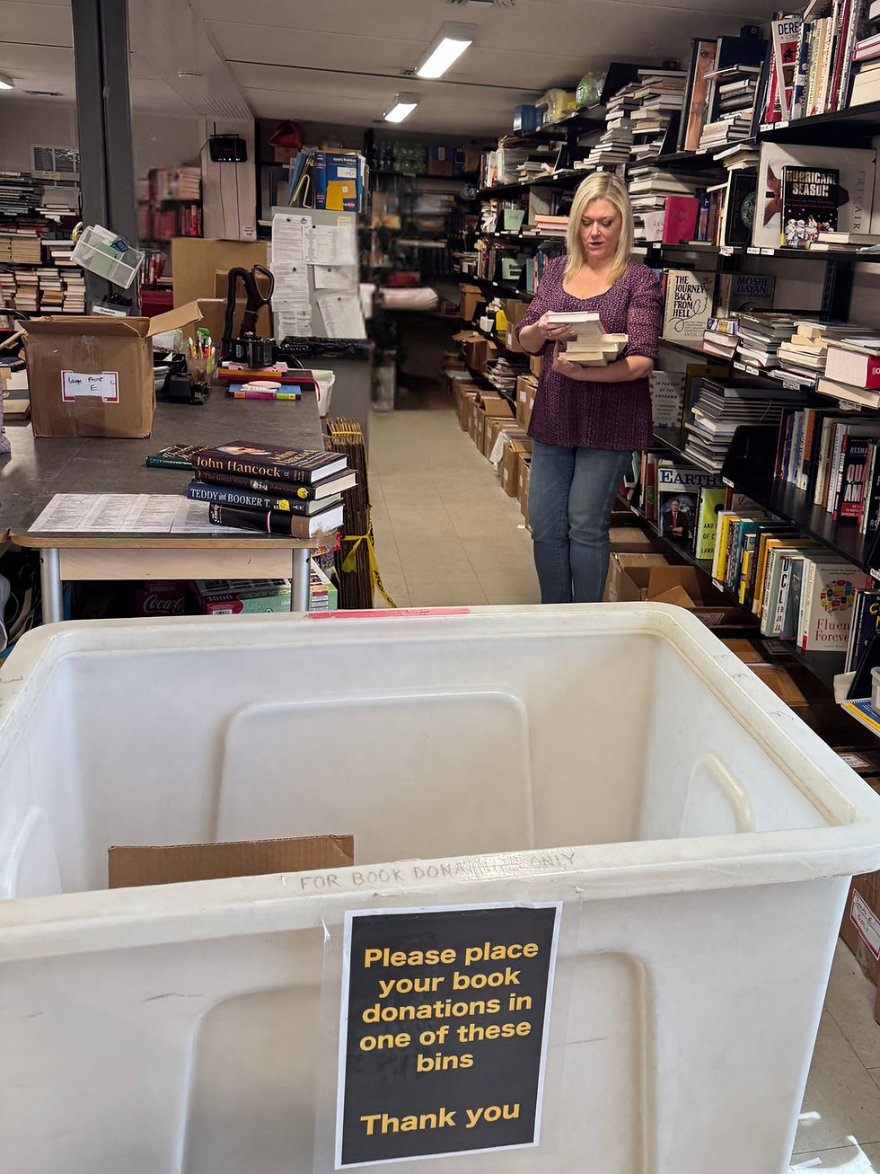 Westport Book Shop volunteer holding books to sort, near a large bin with a sign: "Please place your book donations in one of these bins. Thank you."