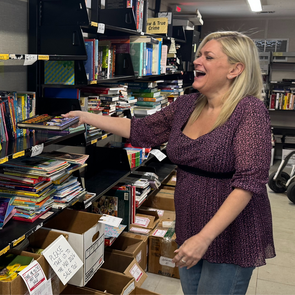 Woman in a purple top smiles while arranging books on shelves. Boxes and sale signs are visible.