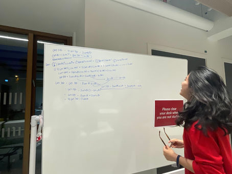 Woman in red sweater holds glasses, looking at math equations on a whiteboard. Red sign reads "Please clear your desk when you are not studying."
