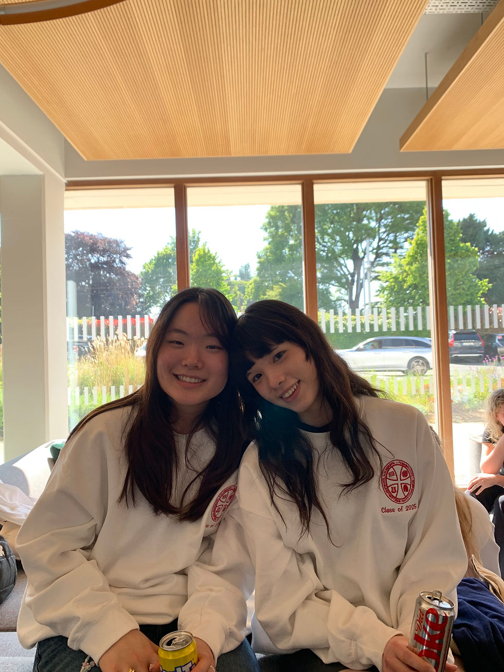 Two friends in white "Class of 2025" sweatshirts smiling, each holding a drink can. Bright room with large windows showing trees outside.