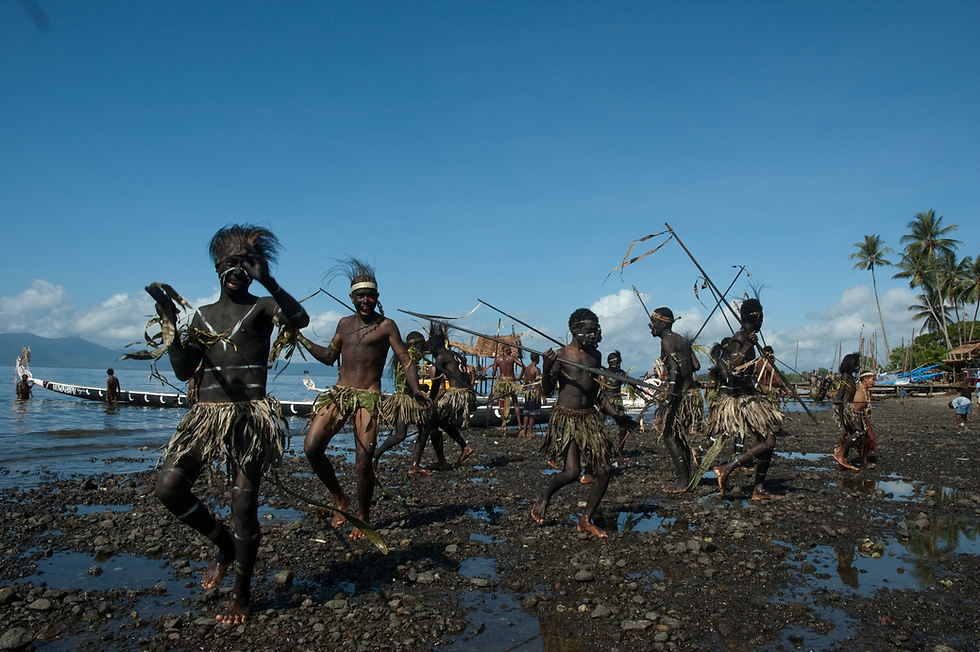 A group of men in traditional dress move together along the shoreline in Milne Bay, with canoes and the sea behind them.