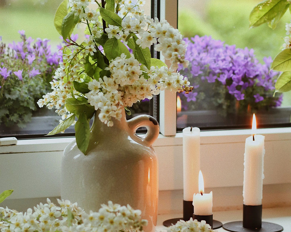 White flowers in a ceramic jug sit on a windowsill with lit candles. Purple flowers bloom outside, creating a serene, cozy atmosphere.