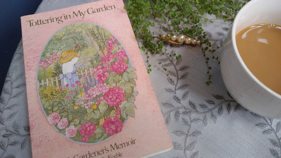 Book titled "Tottering in My Garden" on a tablecloth with a cup of coffee, a plant, and a decorative bird figurine nearby.