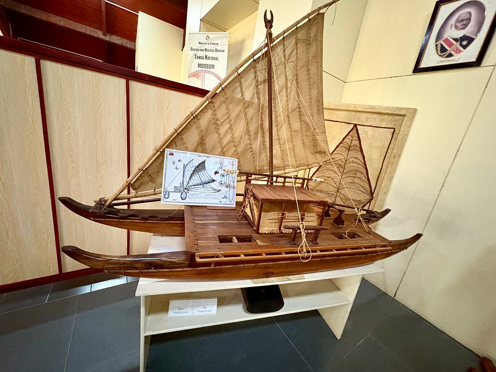 Wooden double-hulled sailing canoe model with tan sail on display in Tonga National Museum, with exhibit sign and framed portrait.