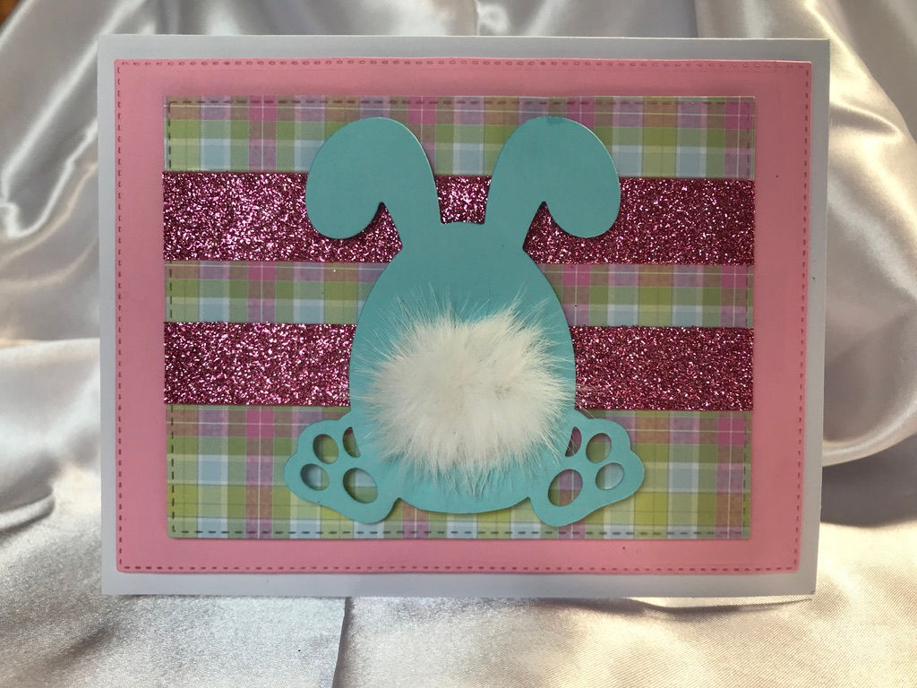 The back of a large blue bunny is the focal point of this card with his fluffy white bottom.  He sits on a pastel, plaid, stitched die cut on top of a pink die cut, on a white card.  Embellished with glittery pink ribbon.
