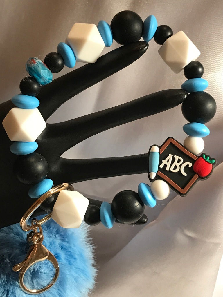 Beaded wristlet with white, black, and blue silicone beads in different shapes and sizes.  One large silicone bead that is a chalkboard with ABC written on it.  A large gold keychain with makes for easy access and a big blue pom pom finishes out the look.