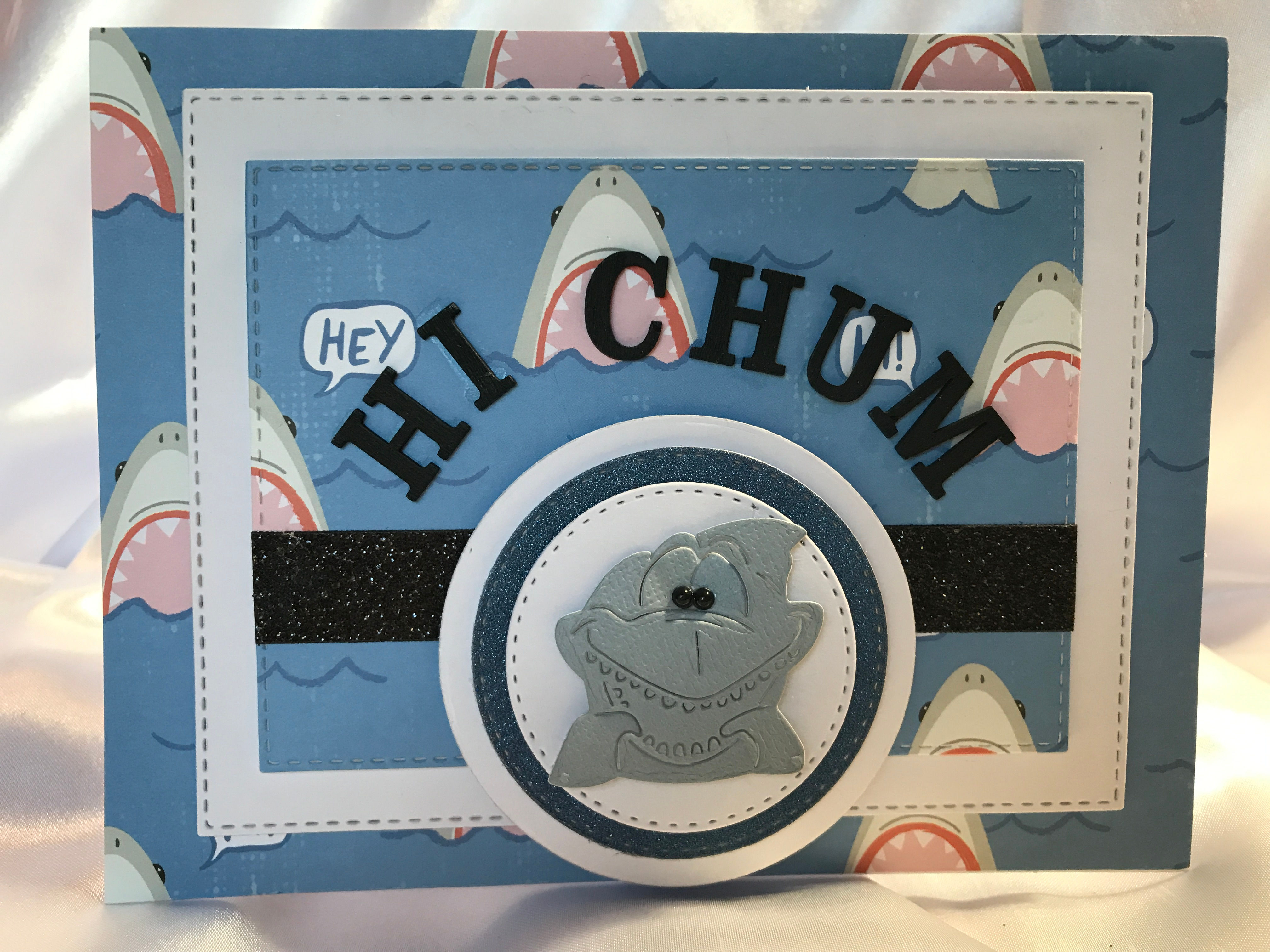This shark is too cute and silly to be scary. He is at the center bottom of this card in grey with black gem eyes. Just his face and fins are showing. The works "Hi Chum" in black wooden letters curve around the top of the die cut circles he is on, they rotate between white and a blue color that coordinates with the background of wide open shark mouths in a blue sea. To add some sparkle there is a small glittery black ribbon embellishment.