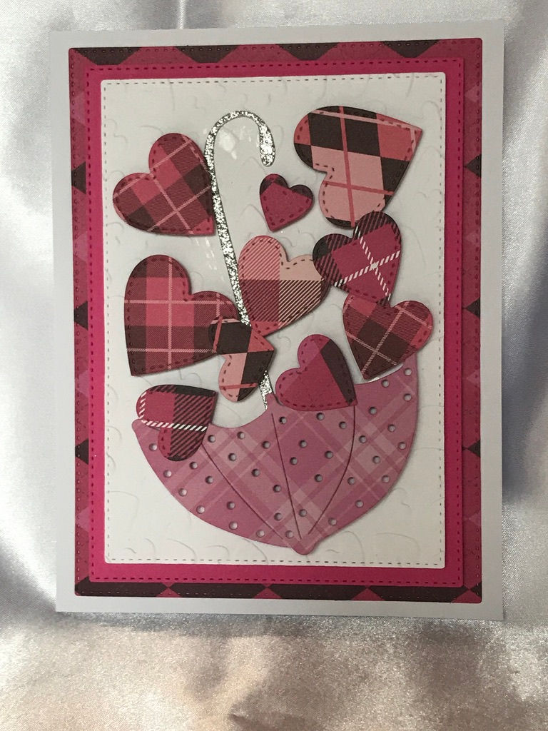 Pink upside down umbrella with glittery silver handle set upside down on a white heart embossed bed.  The umbrella sits upside down to collect all of the falling hearts in varies shades of pink and white plaid prints.    Behind the white is a pink and then a plaid pink layer of die cut card stock on a white card.