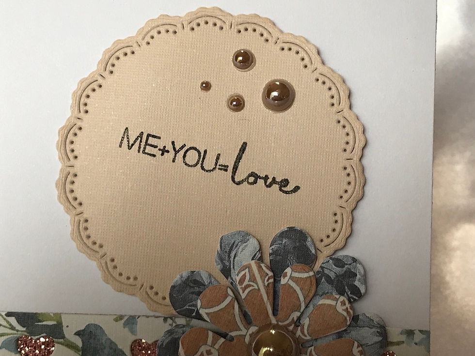Thumbnail: Gorgeous scalloped blue, green, and cream floral border on both the top and bottom of the card, each with a glittery coppery brown ribbon.  On top a row of three die cut flowers with wooden heart buttons on top of each.  All set on a cream and brown multi lined checkered card.