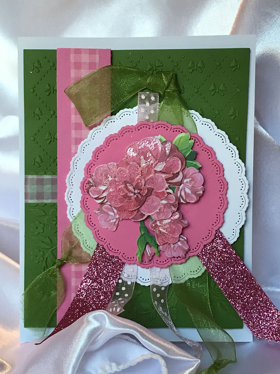Beautifully embossed green card stock with bows and cross stitch diamonds.  Pink and white checkered washi tape with on a pink card stock.  In the center, pink and white scalloped die cut circles with pink polka dot organza ribbon and green organza ribbon tied in a knotted end underneath a bed of pink flowers with green leaves.