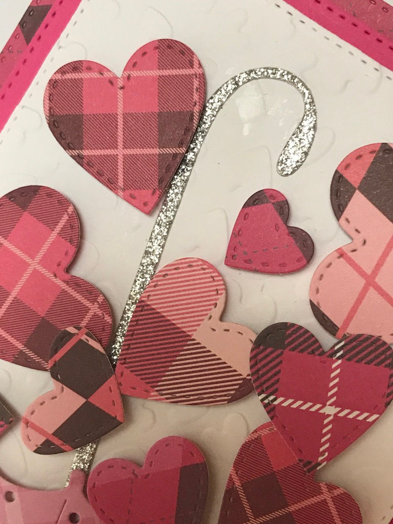Pink upside down umbrella with glittery silver handle set upside down on a white heart embossed bed.  The umbrella sits upside down to collect all of the falling hearts in varies shades of pink and white plaid prints.    Behind the white is a pink and then a plaid pink layer of die cut card stock on a white card.