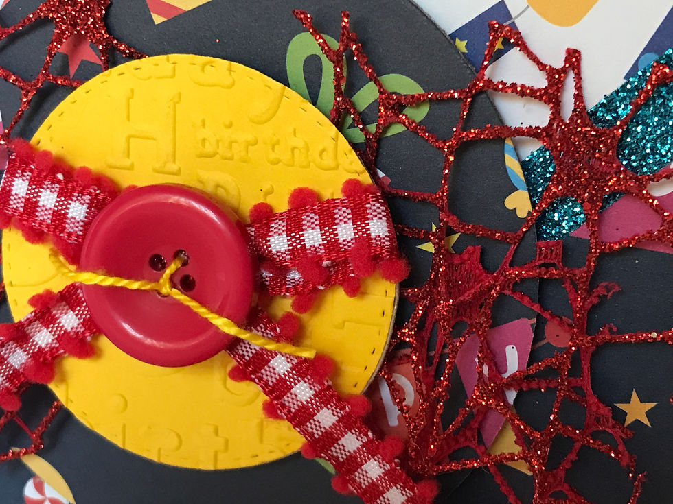 Primary colors in assorted birthday prints make up the background with an embossed yellow center, a big red button, and red and white checkered ribbon under the button.  The embossed yellow circle says Happy Birthday in several fonts, in different directions.  Embellished with a glittery red mesh and glittery blue ribbon diagonal borders on either side.
