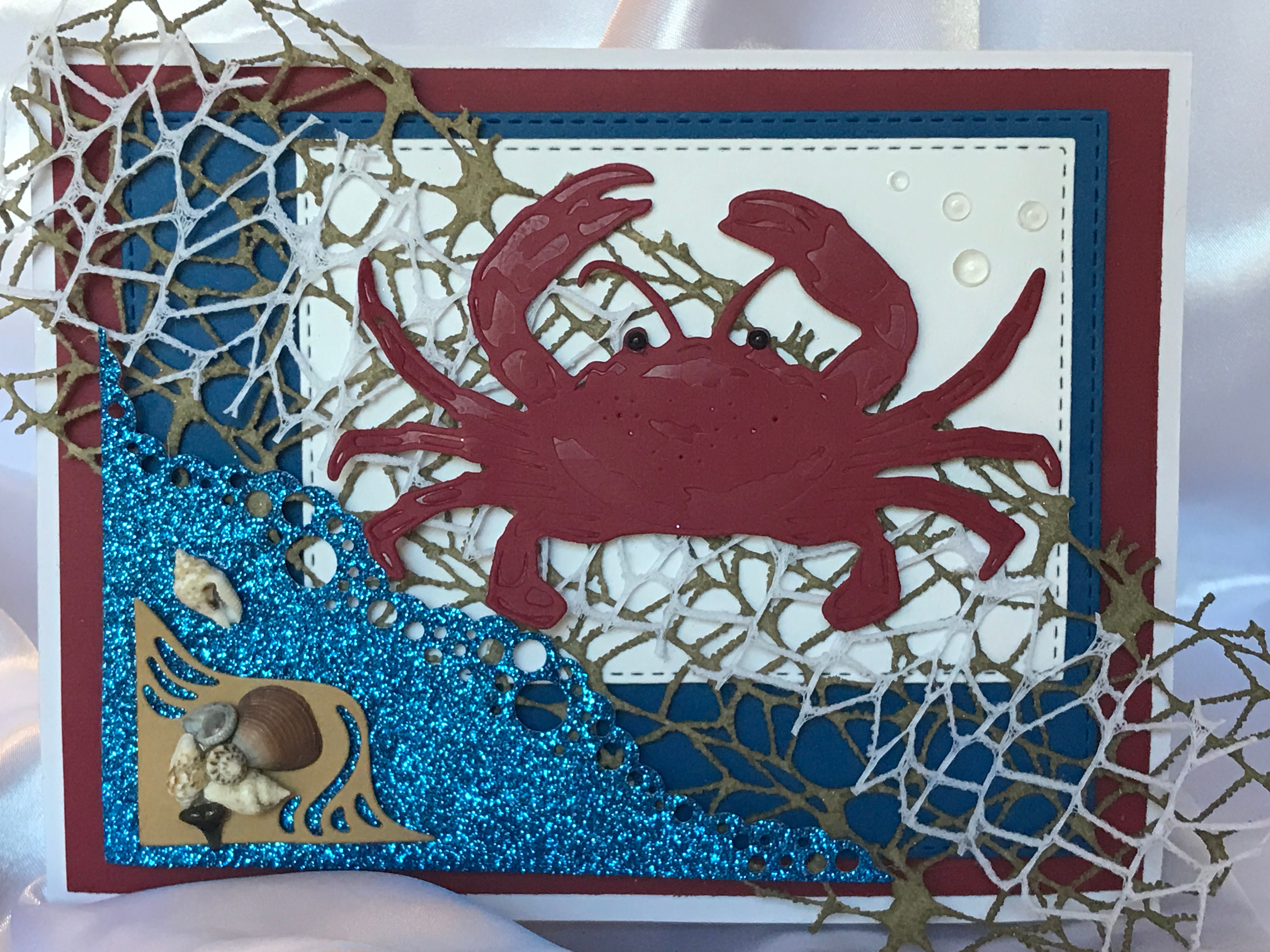 Vibrant die-cut red crab with black acrylic eyes on white card with intricate white and tan underlay. Glittery blue ocean wave die cut with real sea shells and a sharks tooth from Mickler's Beach in St. Augustine, Fl.