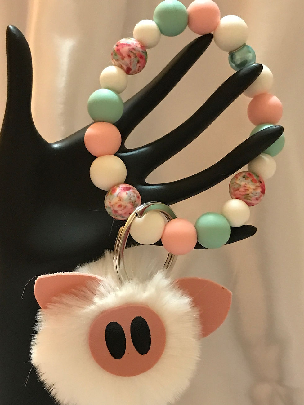 Teal, white, and pink silicone beads in assorted sizes, with three printed pink beads in a floral print with small pink pigs.  A center shinny teal bead with a large key ring and a fluffy white pom pom pig.   He is too cute.