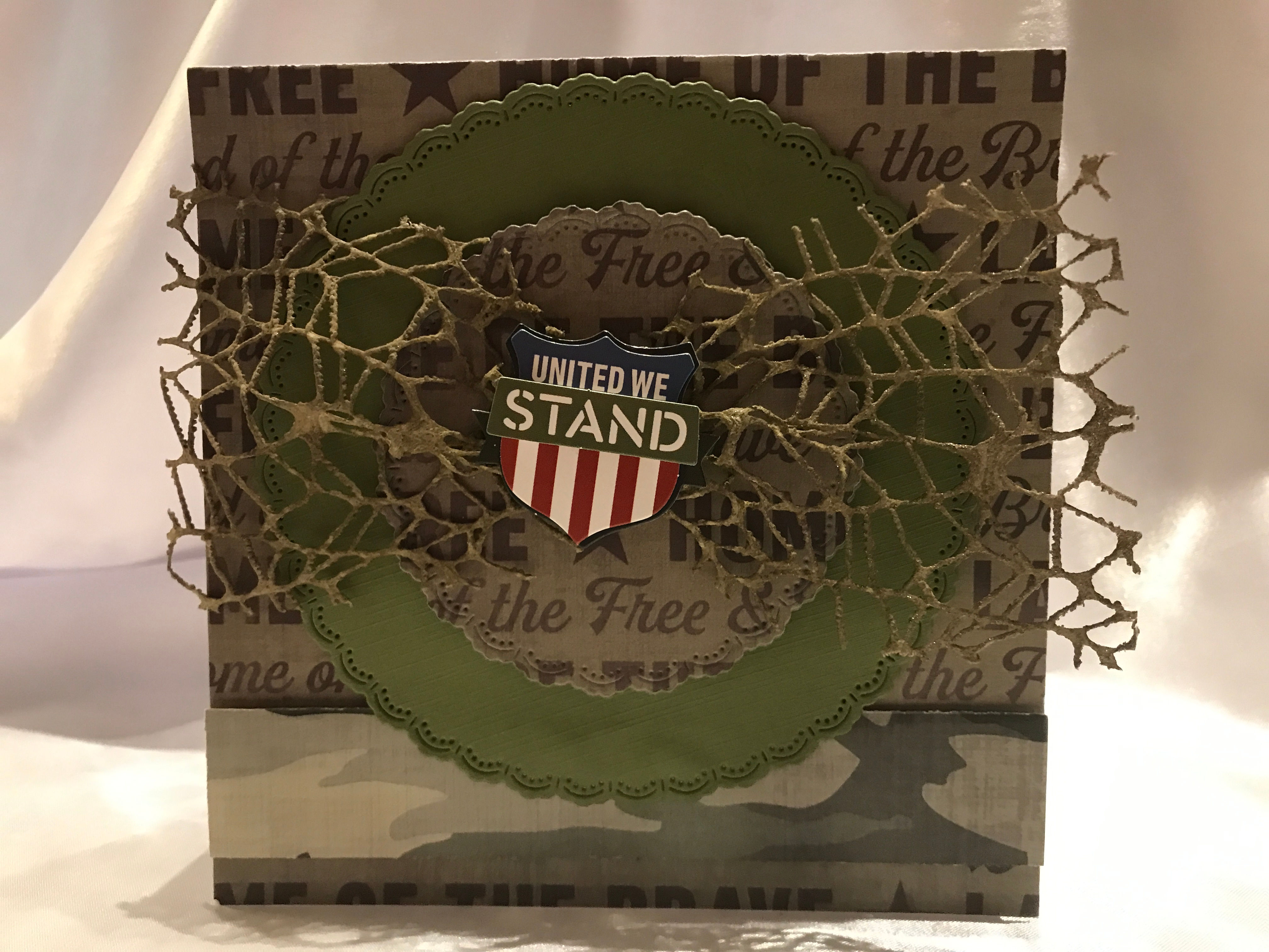 Military themed card, 'UNITED WE STAND' emblem. with olive green scalloped circle with Home of the Free background. Tan mesh embellishment helps make the paper pop.