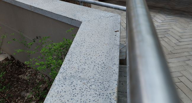 Corner detail where a stainless-steel handrail bracket anchors to a precast planter coping.