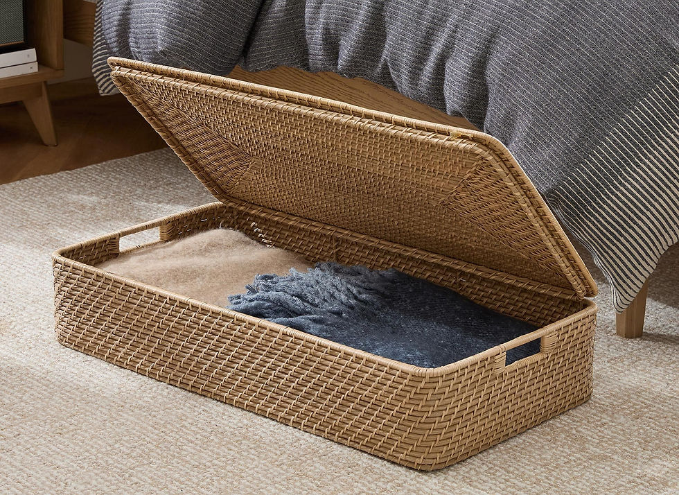 Open wicker under the bed storage box on a beige carpet, containing blue and brown blankets. A striped gray bedspread is partially visible in the background.