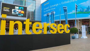 Large yellow "Intersec" sign in front of a building. Blue banners and screens display event messages. People in the background.