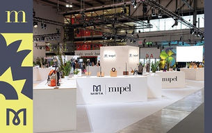 Trade show booth for Mirta and Mipel, featuring handbags on white displays. Bright, modern setting with plants and overhead lights.