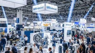 Crowded industrial trade show with people networking among machinery displays. Overhead signs read "GIFA NEWCAST." Brightly lit hall.