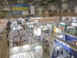 Busy trade show with numerous booths in a large hall. Signs include "KOREA METAL WEEK." Visitors explore displays under bright lights.