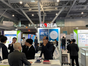 People in business attire converse at a technology expo with a "Korea" sign overhead. Booths display charts and materials, creating a busy atmosphere.