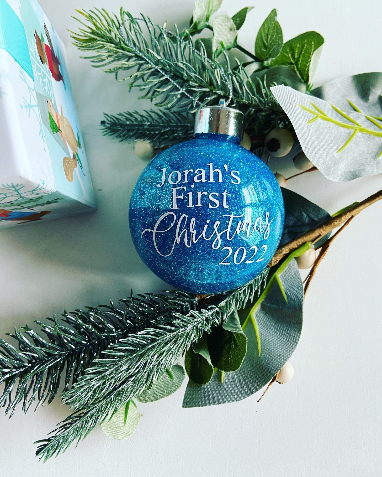 Blue ornament reading "Jorah's First Christmas 2022" nestled in greenery, Christmas theme.