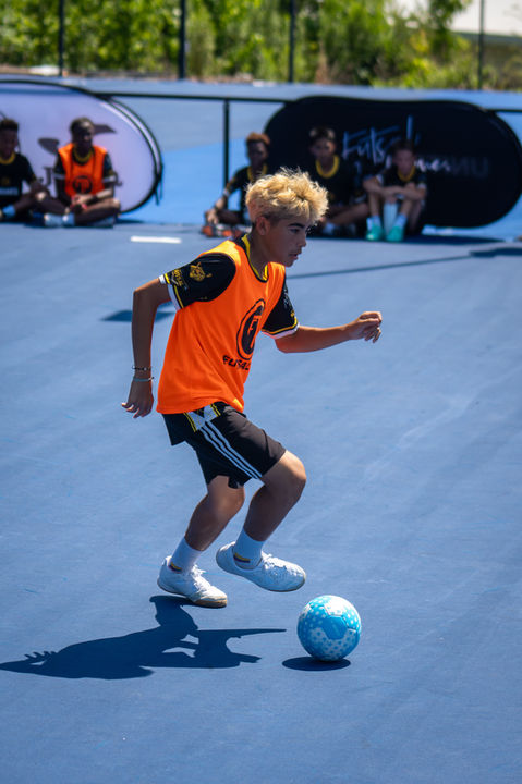 Young boy in orange vest dribbling a soccer ball on a blue court
