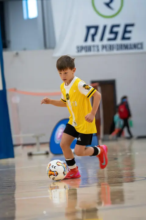 Young boy dribbling soccer ball, RISE SPORTS PERFORMANCE banner, indoor training.