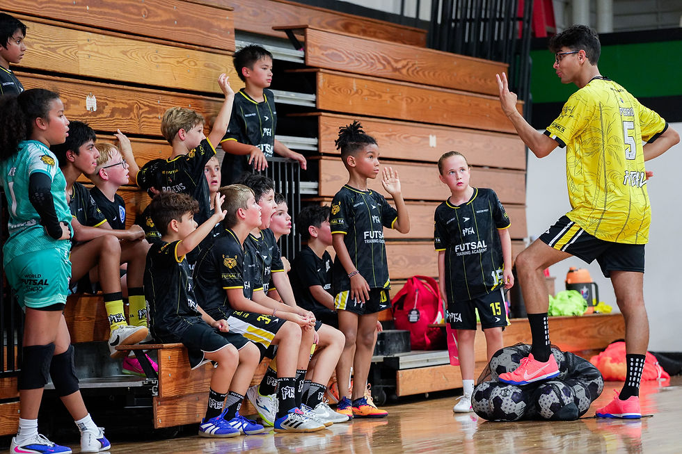 Coach in yellow futsal jersey leads kids in black uniforms, all raising hands in a gym by wooden bleachers.