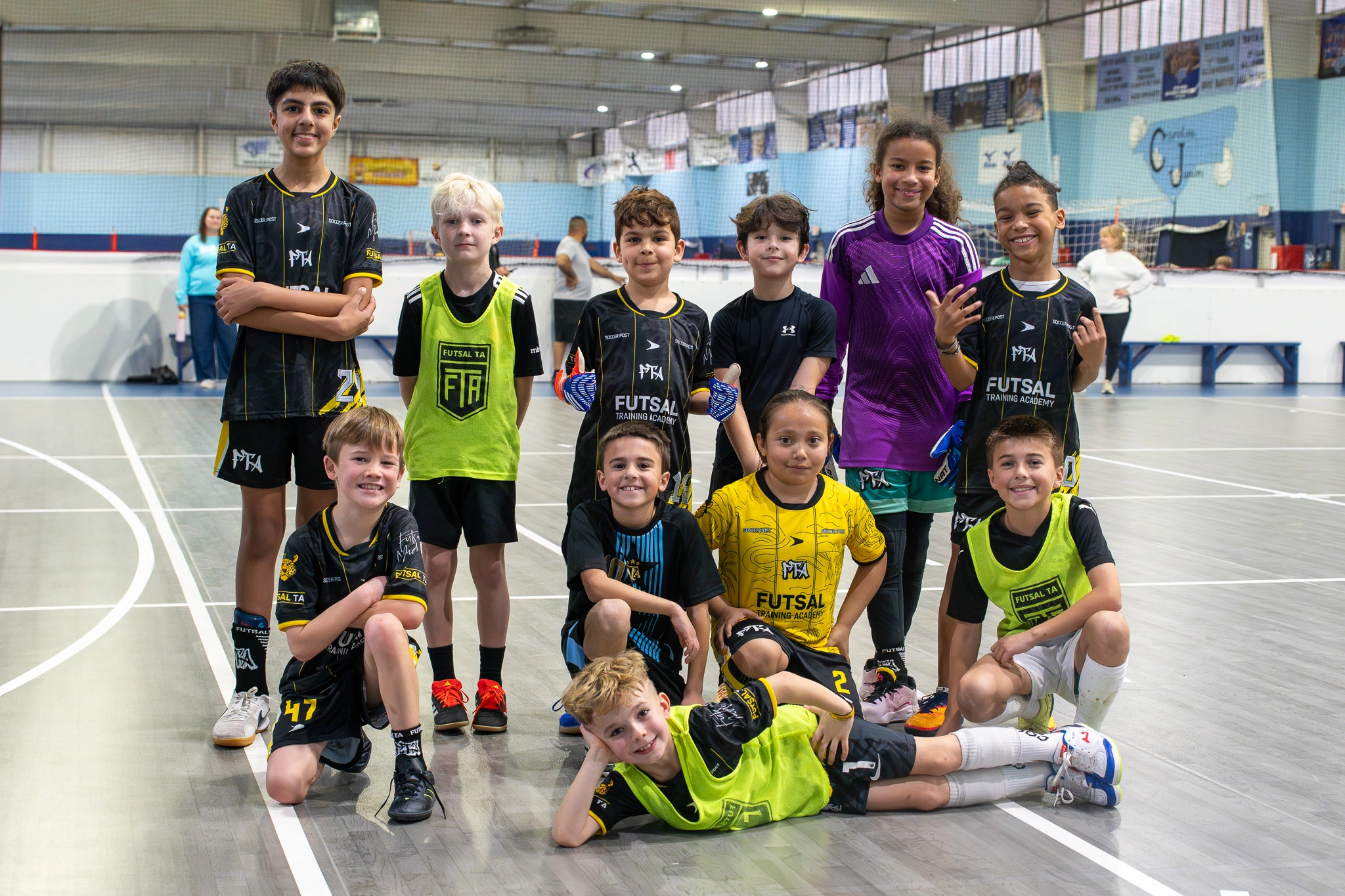 Group of youth futsal players posing together in black and yellow kits inside indoor court facility.