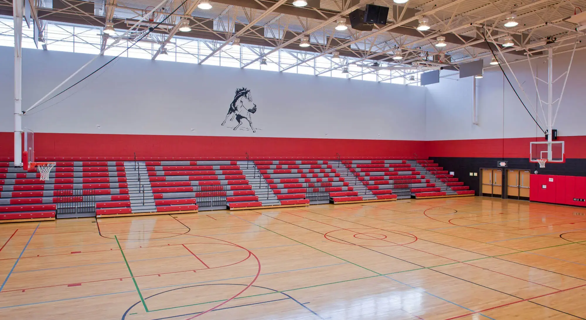 Empty school gym with red bleachers, basketball hoops, and a horse logo on the wall under bright lights