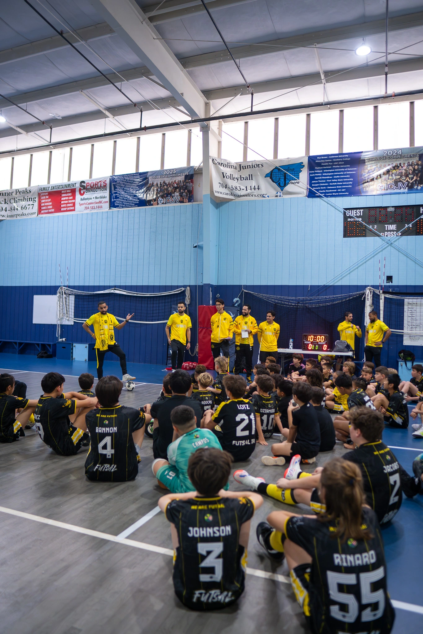 Futsal coach addressing large group of academy players during team talk inside indoor facility.