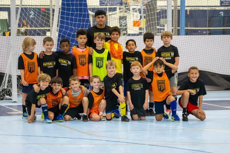 Youth players wearing black and orange FTA gear smiling on indoor futsal court with goal behind them