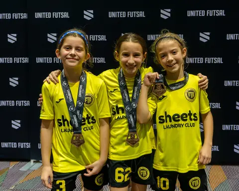 Three young futsal girls smiling with medals and trophies in front of United Futsal backdrop