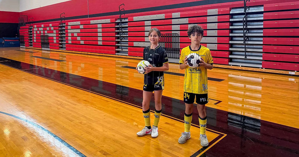 Two youth futsal players in black and yellow uniforms hold balls in a red-and-black gym, with FUTSAL TRAINING ACADEMY on jerseys.