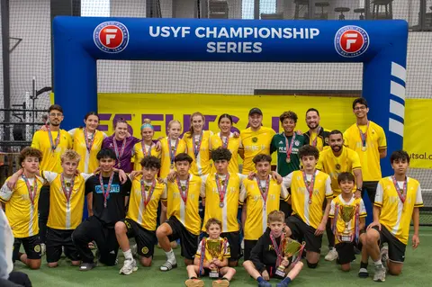 A group of boys posing for a photo under a USYF Banner. Half standing in yellow and half sitting in Black