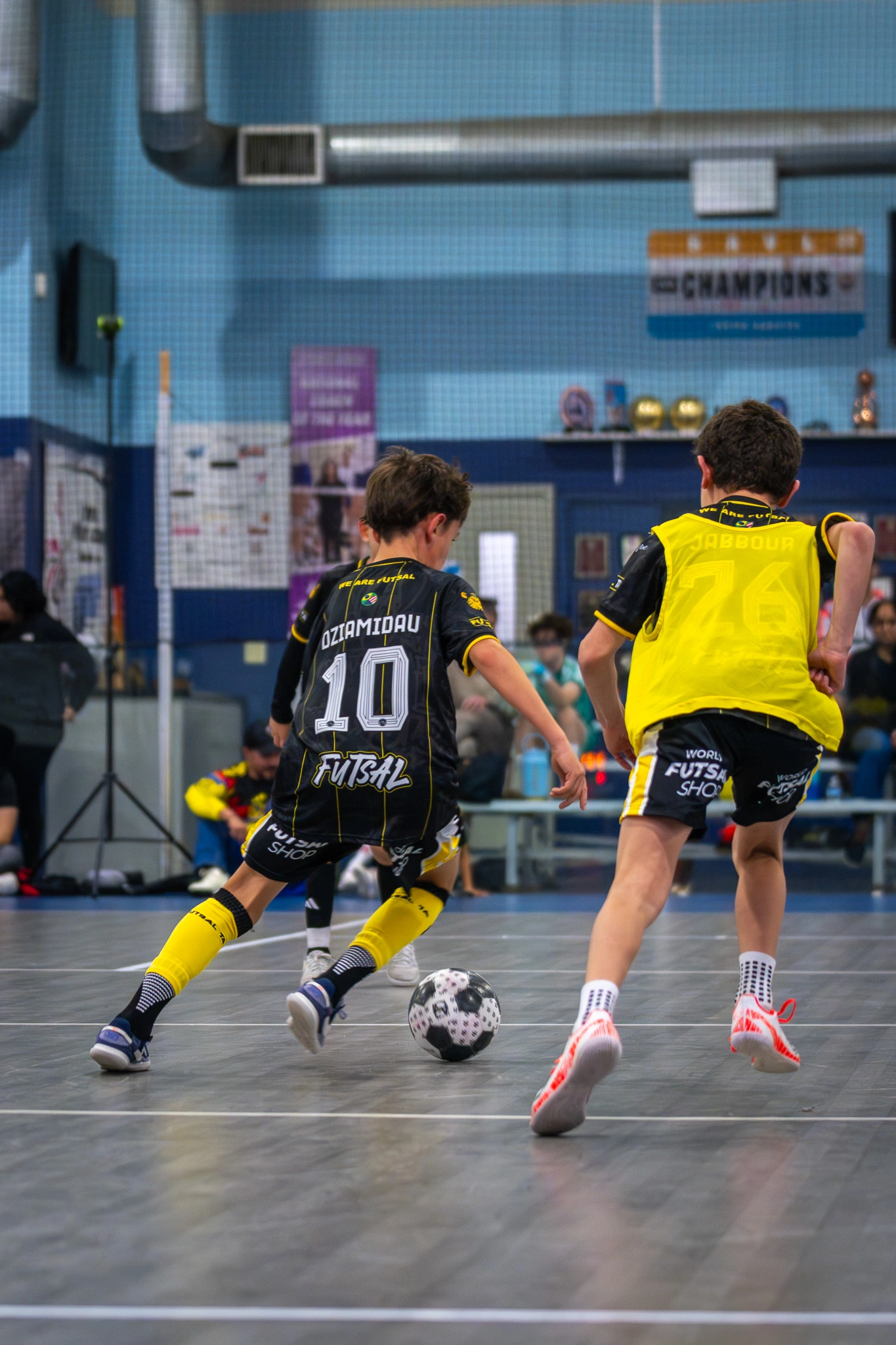 Two youth futsal players competing for the ball in black and yellow FTA uniforms.