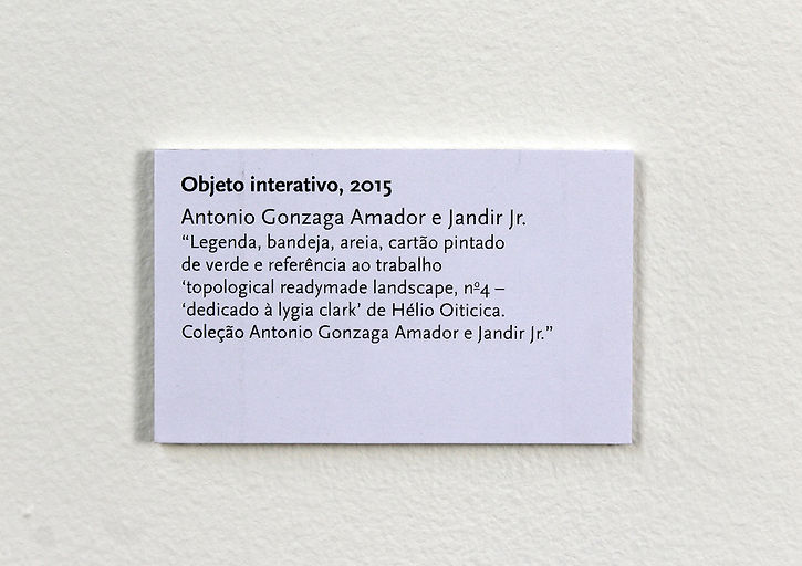 Amador e Jr. Segurança Patrimonial Ltda. Performance Objeto interativo. Legenda de parede da obra que acompanhou a performance. Nela, está escrito: Objeto interativo, 2015. Antonio Gonzaga Amador e Jandir Jr. Legenda, bandeja, areia, cartão pintado de verde e referência ao trabalho Topological readymade landscape, número 4 - dedicado à Lygia Clark, de Hélio Oiticica. Coleção Antonio Gonzaga Amador e Jandir Jr.