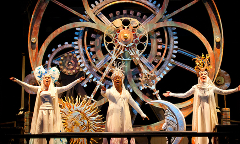 Three women stand on a balcony in elaborate white dresses, robes, and headpieces. Behind them stands a massive clock with exposed gears.