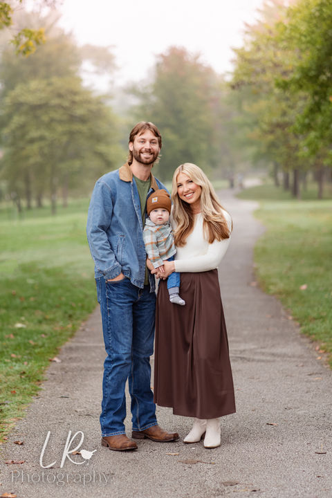Family cuddling by Lake Taneycomo at Branson Landing — dad in denim jacket, mom in white sweater and brown skirt, holding baby in brown hat and plaid shirt surrounded by fall fog and golden trees.