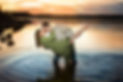 Man in a grey dress shirt dips his fiancée backward for a romantic kiss during sunset in Table Rock Lake, Branson, as she smiles in a flowing green dress, their feet submerged in the calm water.
