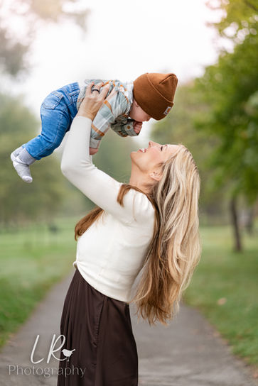 Mom playfully holds baby in the air by Lake Taneycomo at Branson Landing — mom in white sweater and brown skirt, holding baby in brown hat and plaid shirt surrounded by fall fog and golden trees.