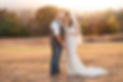 Newlyweds lean their heads together with eyes closed beneath a glowing orange and golden autumn sunset, her veil gently blowing in the breeze, capturing a quiet, tender moment just after saying “I do.”