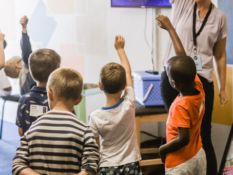 Students raising their hands in an inclusive classroom with a teacher providing support.