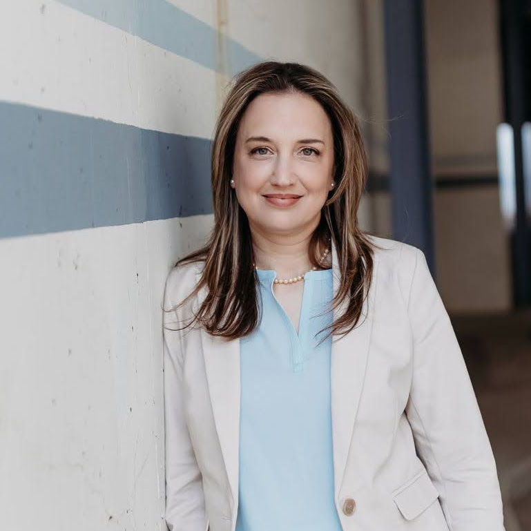 A woman with brown hair wearing a light blue top and white blazer, smiles while leaning against a striped wall.