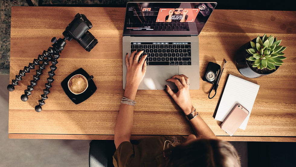 Ein Schreibtisch von oben auf dem eine Kamera mit Equipment, ein Kafee und ein Videoediting-Laptop liegen. Eine Frau bedient das Laptop auf dem sichtbar eine Videoschnittsoftware läuft. Ihre Hände ruhen aktiv über der Tastatur.