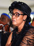 Queen Mother Best Kemigisa seated at a formal outdoor event, wearing a black hat, glasses, and elaborate black lace attire, during the period surrounding King Oyo’s burial and the reported incident involving her being locked in a car.