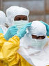 Healthcare workers in yellow hazmat suits and masks adjust white hoods outdoors, blue gloves raised in a tense scene. "Caption" Healthcare workers don protective gear as part of the ICRC Ebola response efforts in Ituri province, DRC.